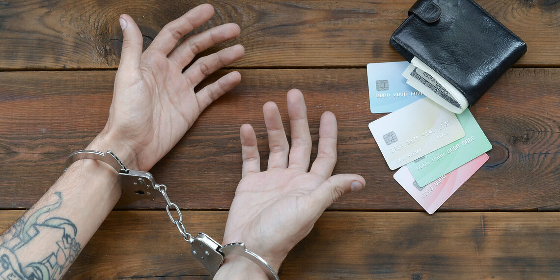 Cuffed hands of tattooed criminal suspect of carding and fake credit cards with cash in purse as evidence for investigation. Above view to old prison wooden table